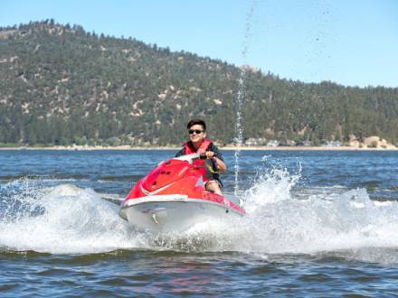 A person in a life jacket rides a red jet ski across a body of water, creating waves in the summer in Big Bear Lake, CA.