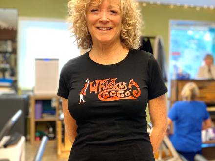 A photo of a Pilates instructor, her hands are behind her back and she has a welcoming smile