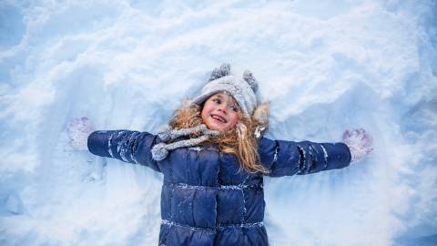 A photo of a little girl in a blue snow jacket making an angel in the fluffy white snow while smiling happily.