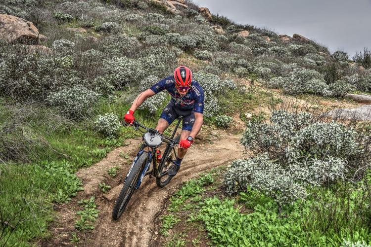 A photo of a male bike rider ascending a hill on it's bike track, his blue shirt has Kenda in bold letters