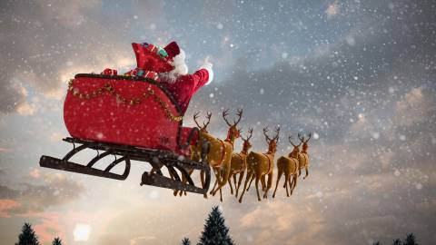 A picture of Santa riding his red sleigh pulled along by his reindeer through the falling snow, the sun beginning to rise.