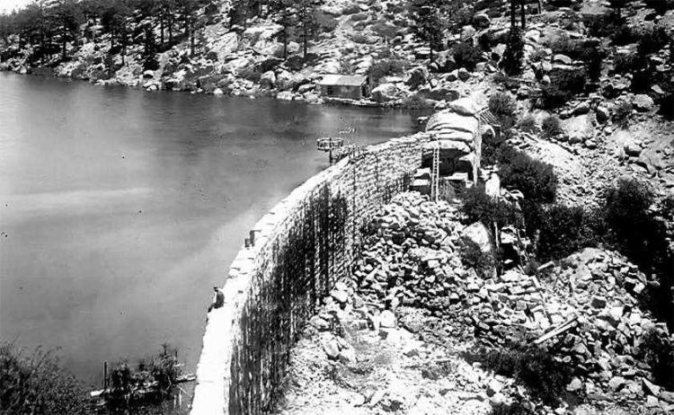 एक आदमी 1911 से पहले बिग बीयर झील के सामने, पहले पत्थर के बिग बीयर झील बांध के किनारे पर बैठा है
