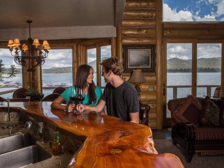 A couple sits at a wooden counter with glasses of wine inside a cabin with a view of Big Bear Lake outside the windows.